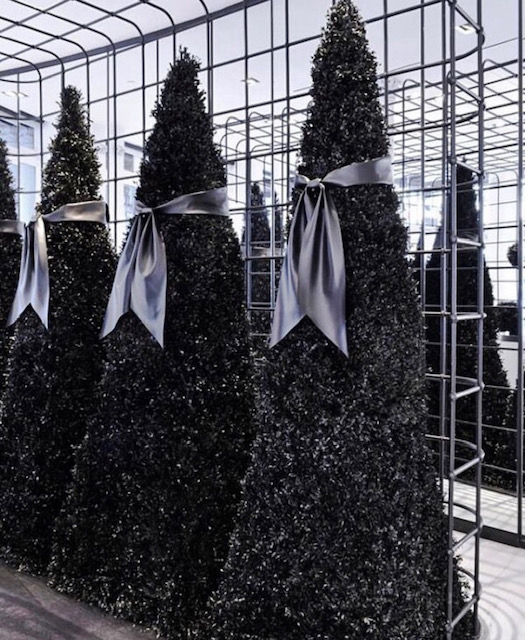 Detail of the Alexander Wang installation — tall black topiaries cinched with grey satin bows against a mirrored grid backdrop